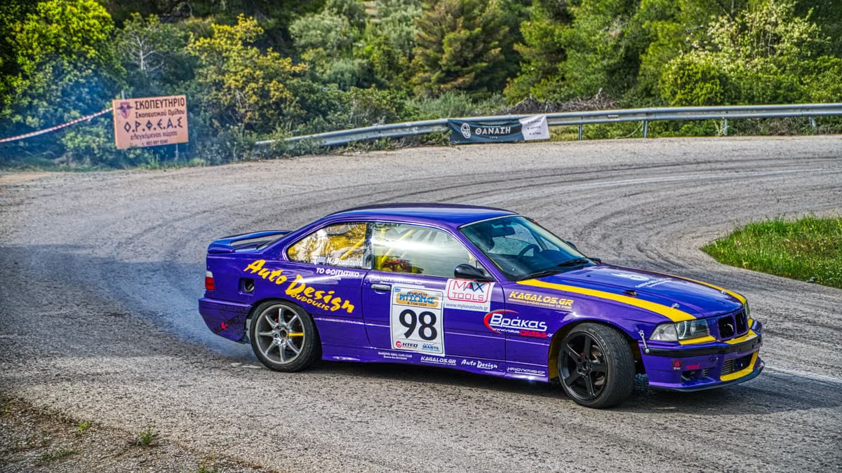A BMW E36 performs a thrilling drift on a winding mountain road during a daytime racing event.